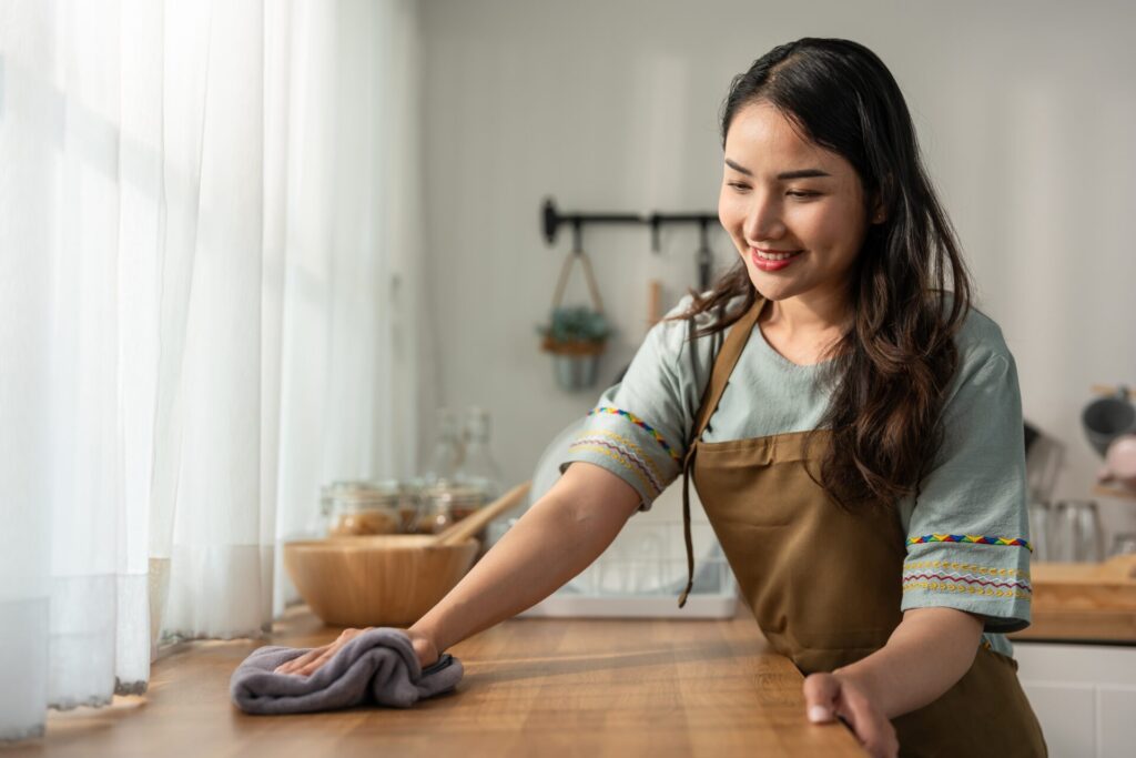 A woman wearing an apron is smiling while cleaning a wooden kitchen counter with a cloth. 