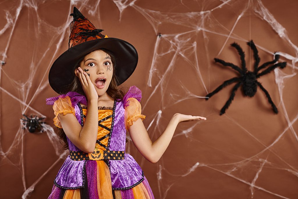 Amazed little girl in a witch costume for Halloween, pointing to a fake very large, black, fuzzy spider on the wall, with large fake spiderwebs.